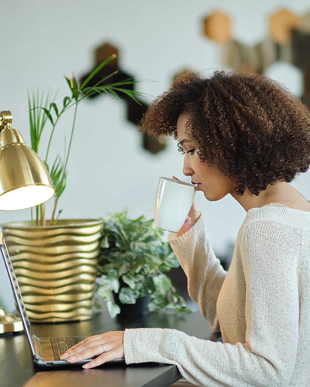 African-American woman working on laptop in front of gold metallic desk lamp in home office decorated with afro-boho glam aesthetic.